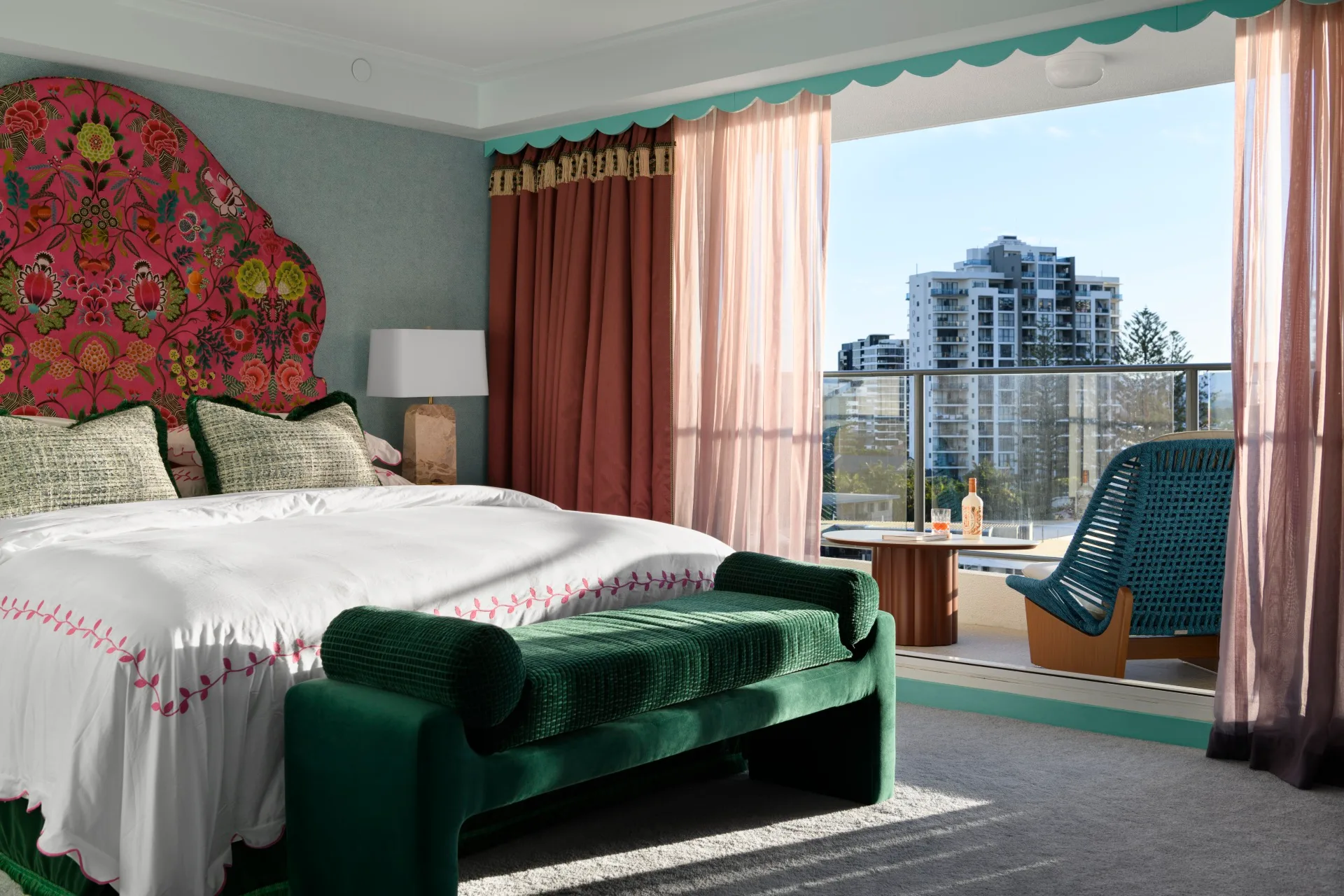 Luxurious hotel room with colorful headboard, elegant drapes, balcony view with city skyline, and modern seating area.