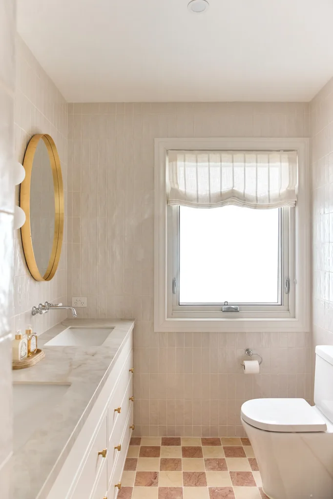 Bright bathroom with a round mirror, marble countertop, and window with light curtains over a toilet and sink.