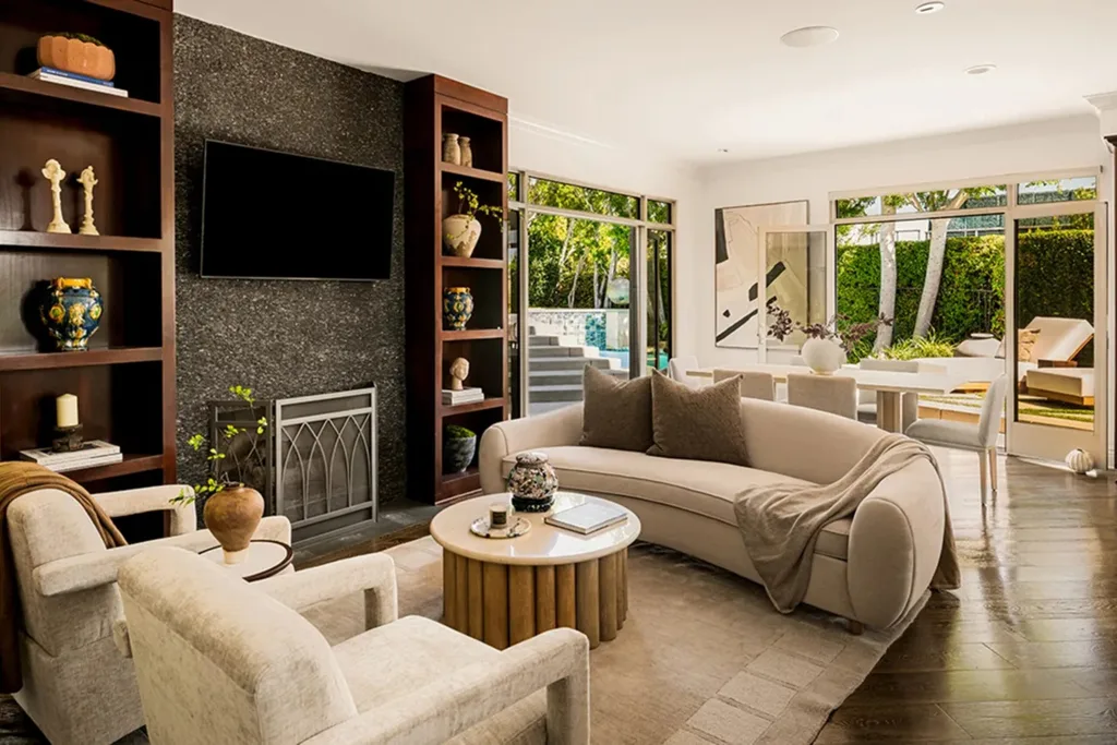 Modern living room with beige furniture, large windows, and wall-mounted TV. Garden view visible outside. Dolly Parton's Hollywood Hills home.
