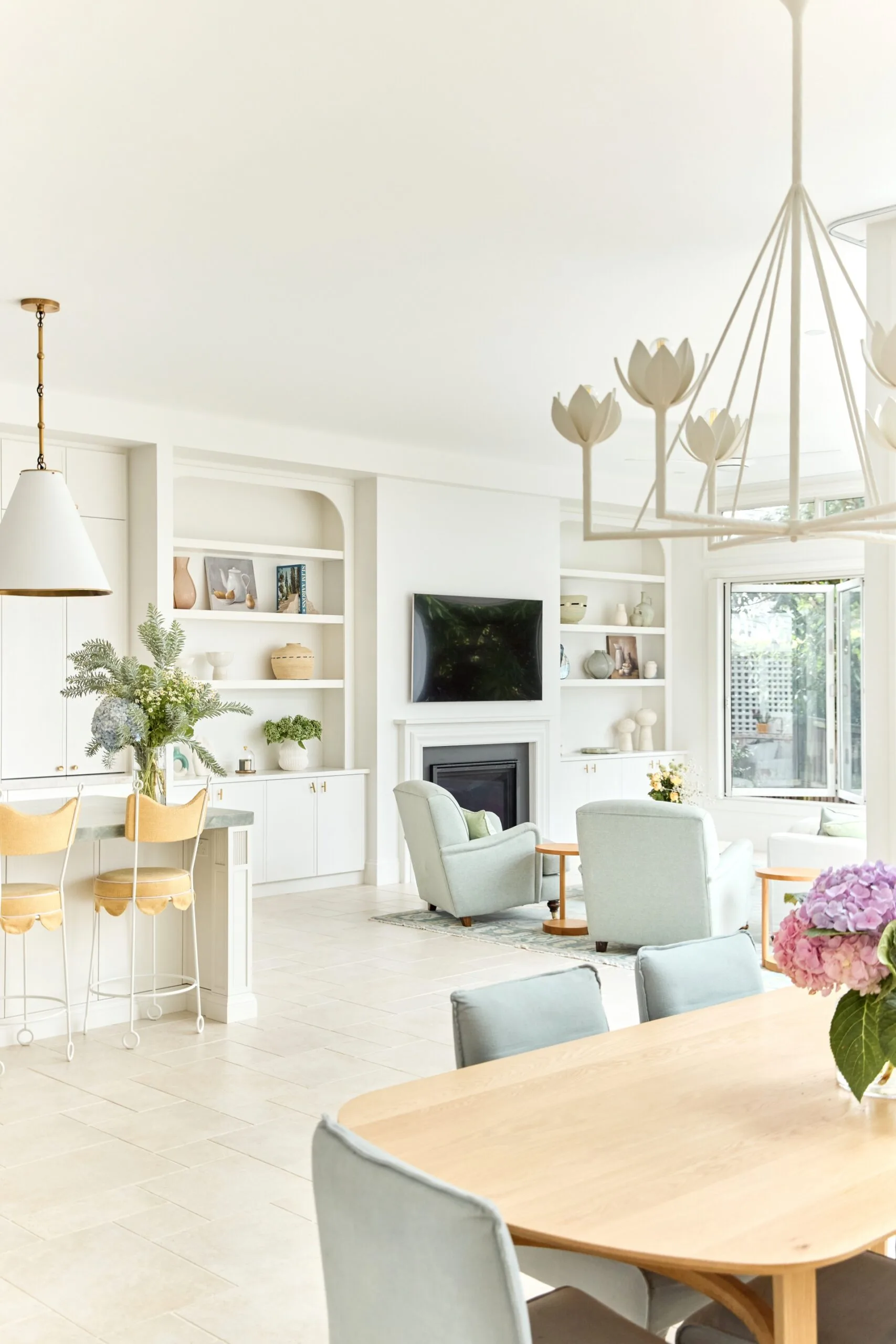Bright, airy living room with light furniture, a fireplace, and open shelves; view of a dining area with flowers.