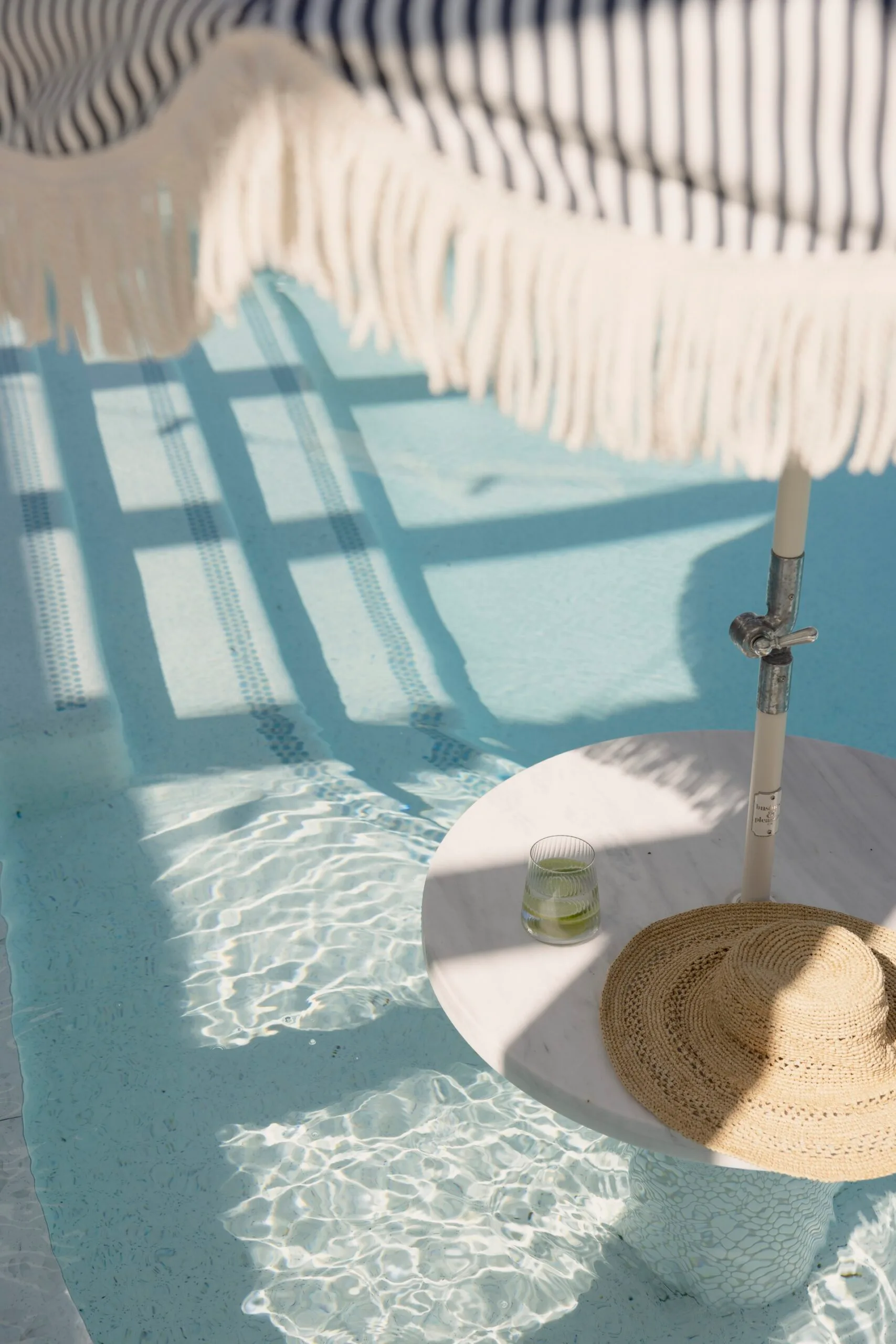 Poolside table with a straw hat and drink, under a striped umbrella casting shadows on the water.