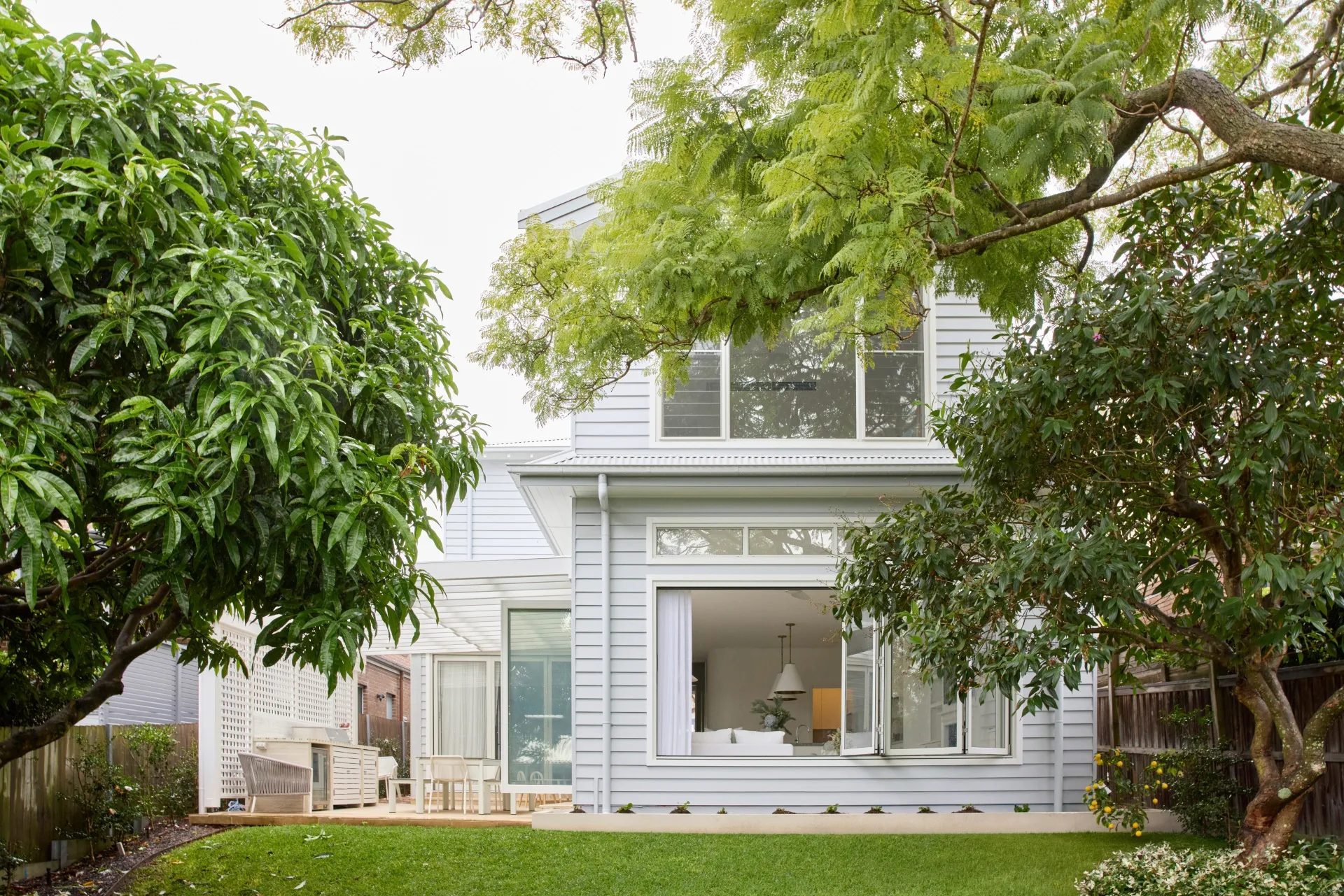 Two-story white house with large windows, surrounded by lush green trees and a grassy yard.