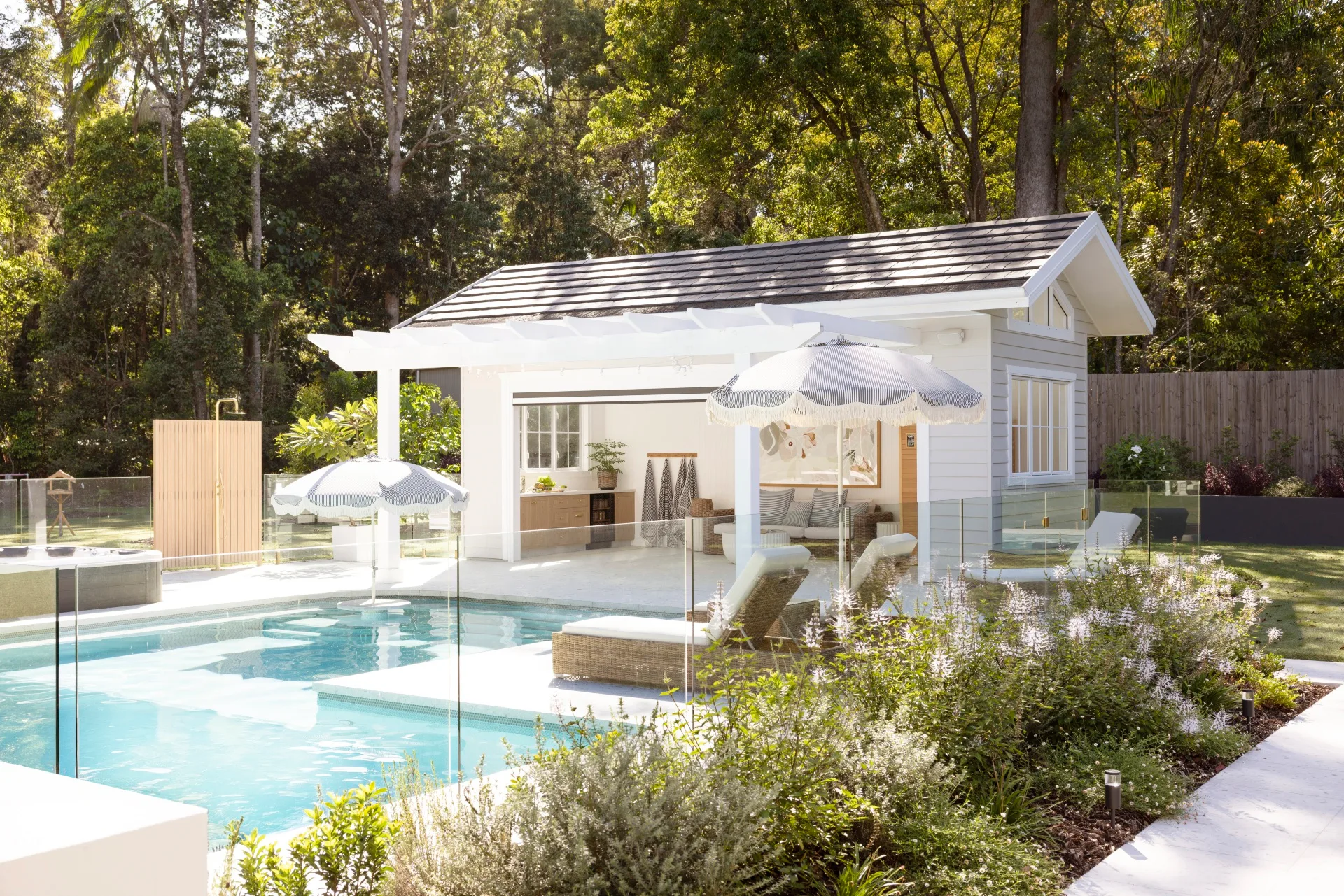 Bright poolside scene with white pergola, loungers, umbrellas, and lush greenery in a tranquil backyard setting.