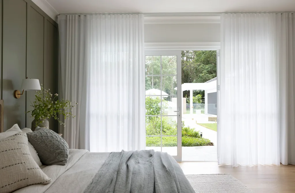 Bedroom with gray pillows and a bedspread, large windows with sheer curtains open to a garden view outside.