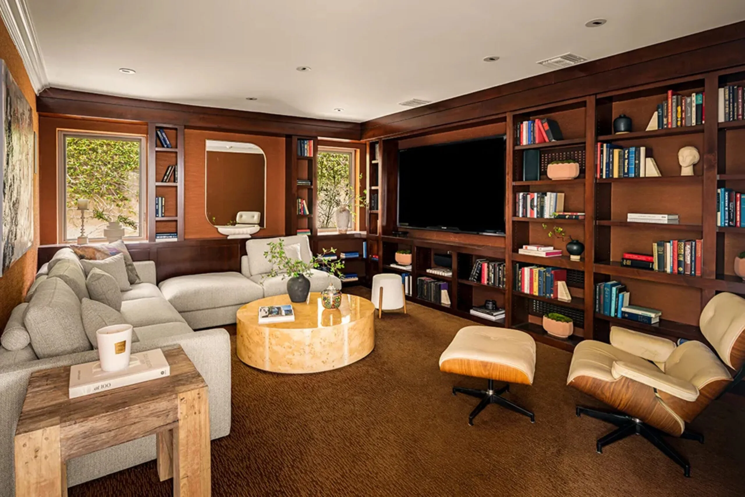 Cozy living room with beige sectional, wooden shelves, books, large TV, round coffee table, and an armchair with ottoman.