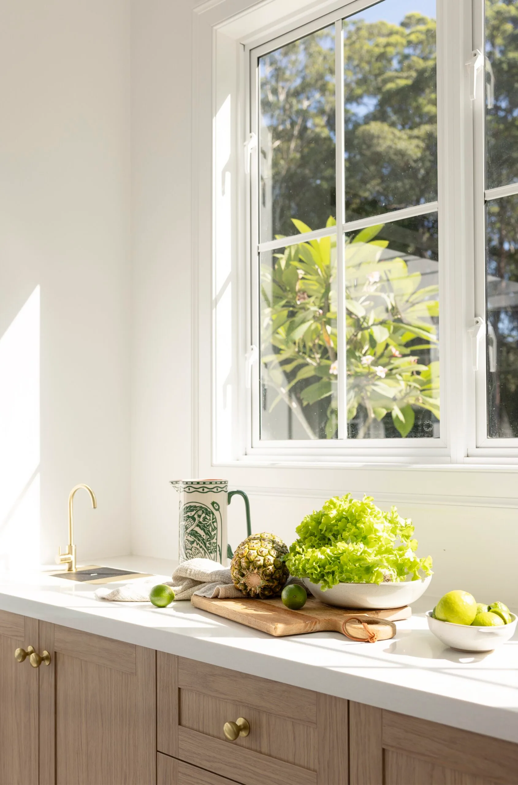 Sunny kitchen with wooden cabinets, a window view, and fresh produce including lettuce, pineapple, and limes on the counter.