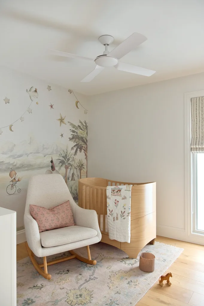 Nursery with wooden crib, rocking chair, floral pillow, whimsical wall mural, and pastel rug.