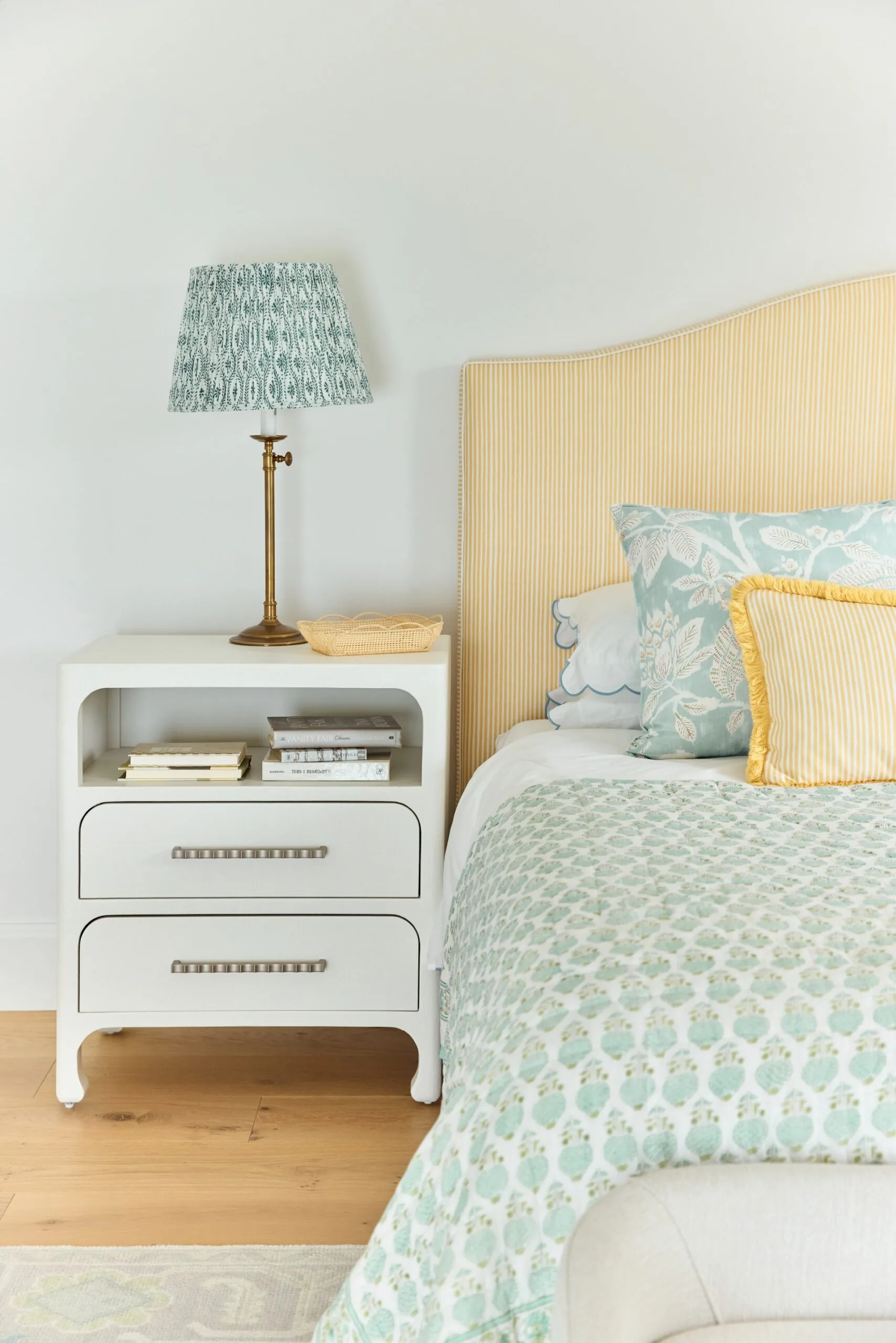 Bedroom with white nightstand, brass lamp, yellow-striped headboard, teal and yellow pillows, quilted bedding.