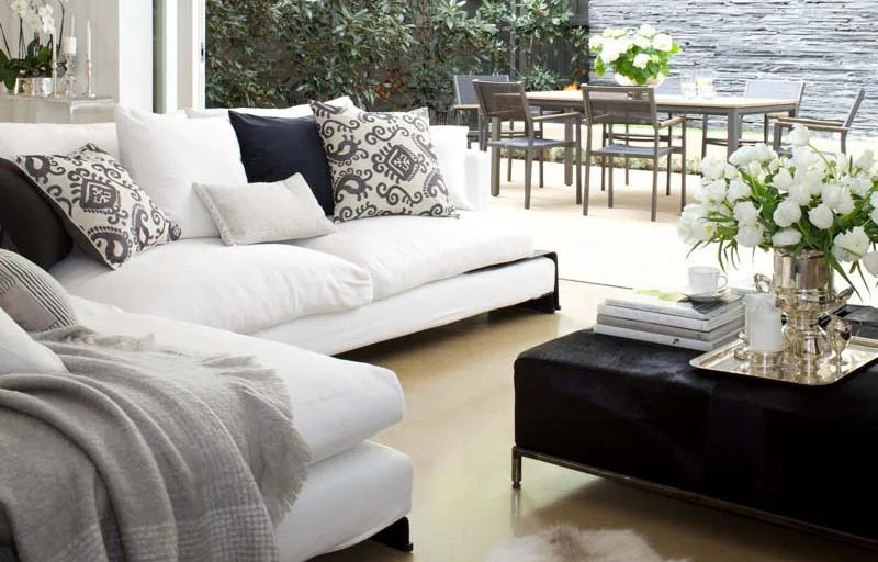 Bright living room with white sofas, patterned cushions, a black ottoman with flowers, and a dining area in the background.