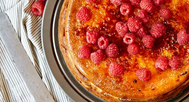 Cheesecake topped with fresh raspberries on a caramelized surface, placed on a striped cloth background.