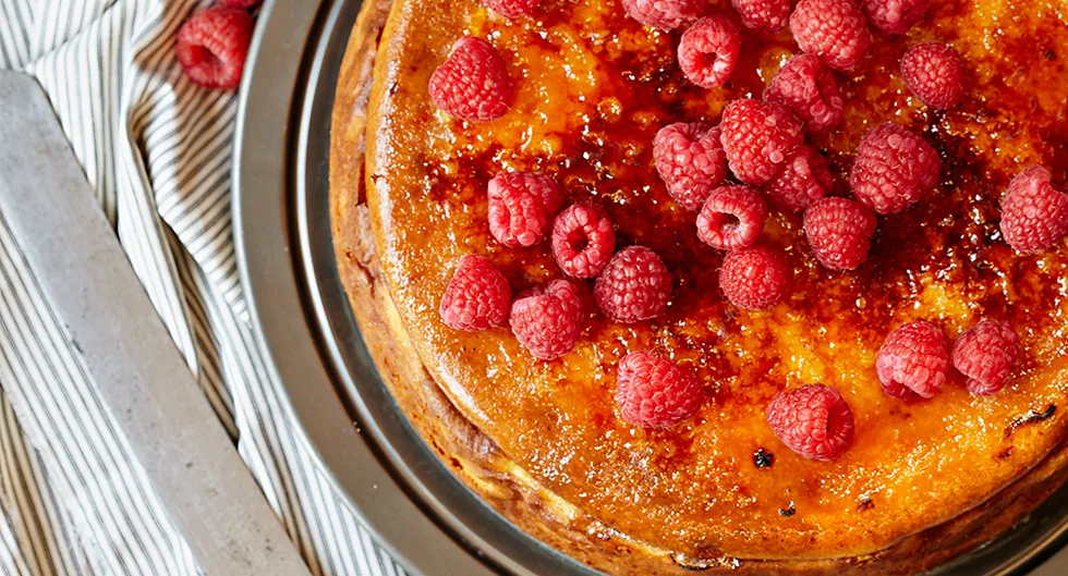 Cheesecake topped with fresh raspberries on a caramelized surface, placed on a striped cloth background.
