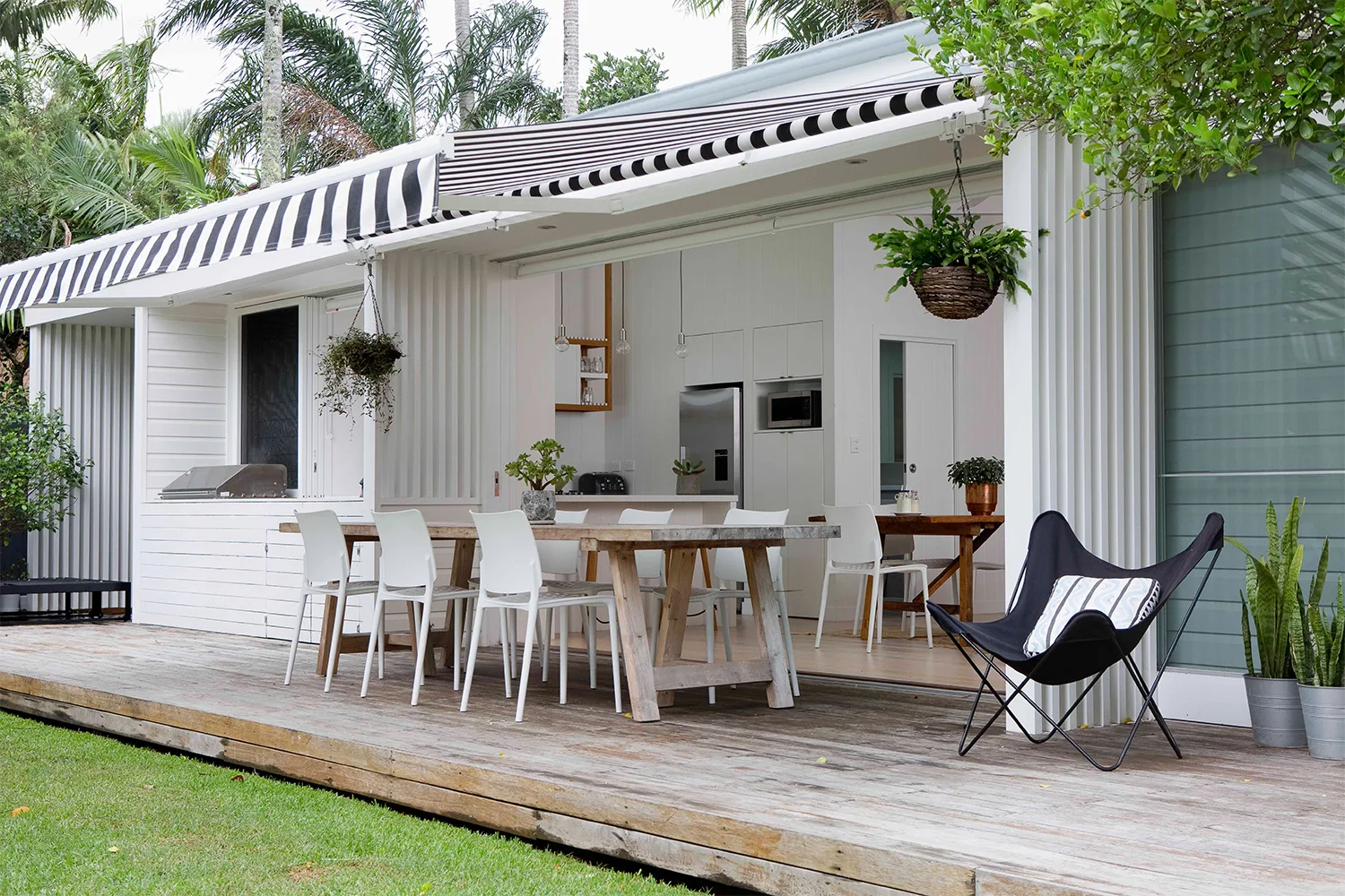 A converted shed granny flat with white painted bricks and a black and white striped fabric awning over a timber deck with dining furniture