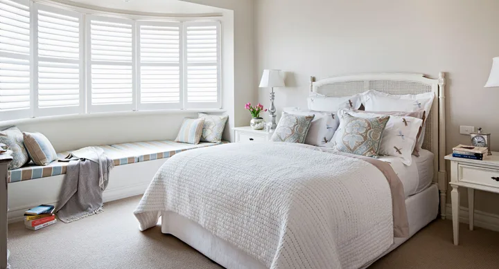 Bedroom with white bedding, a window seat with cushions, books, a table with flowers and lamp, and wooden shutters.