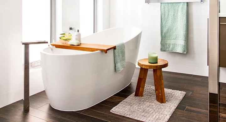 Modern bathroom with a white freestanding bathtub, wooden stool, green towel, and candle on a textured rug.