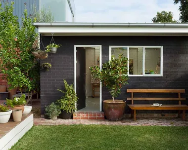 Black brick granny flat with white windows and doors in green backyard.