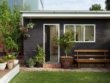 Black brick granny flat with white windows and doors in green backyard.