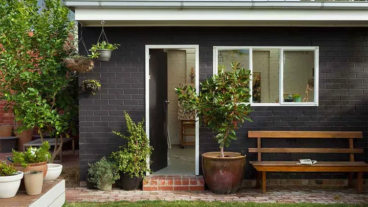 Black brick granny flat with white windows and doors in green backyard.