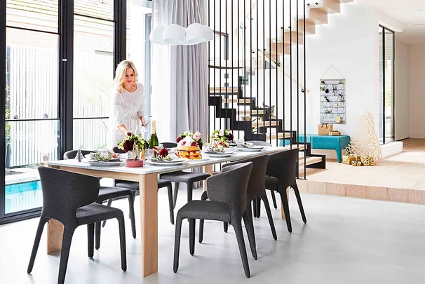 Blonde woman setting a modern dining table with elegant decor in a bright, open living space with a floating staircase.
