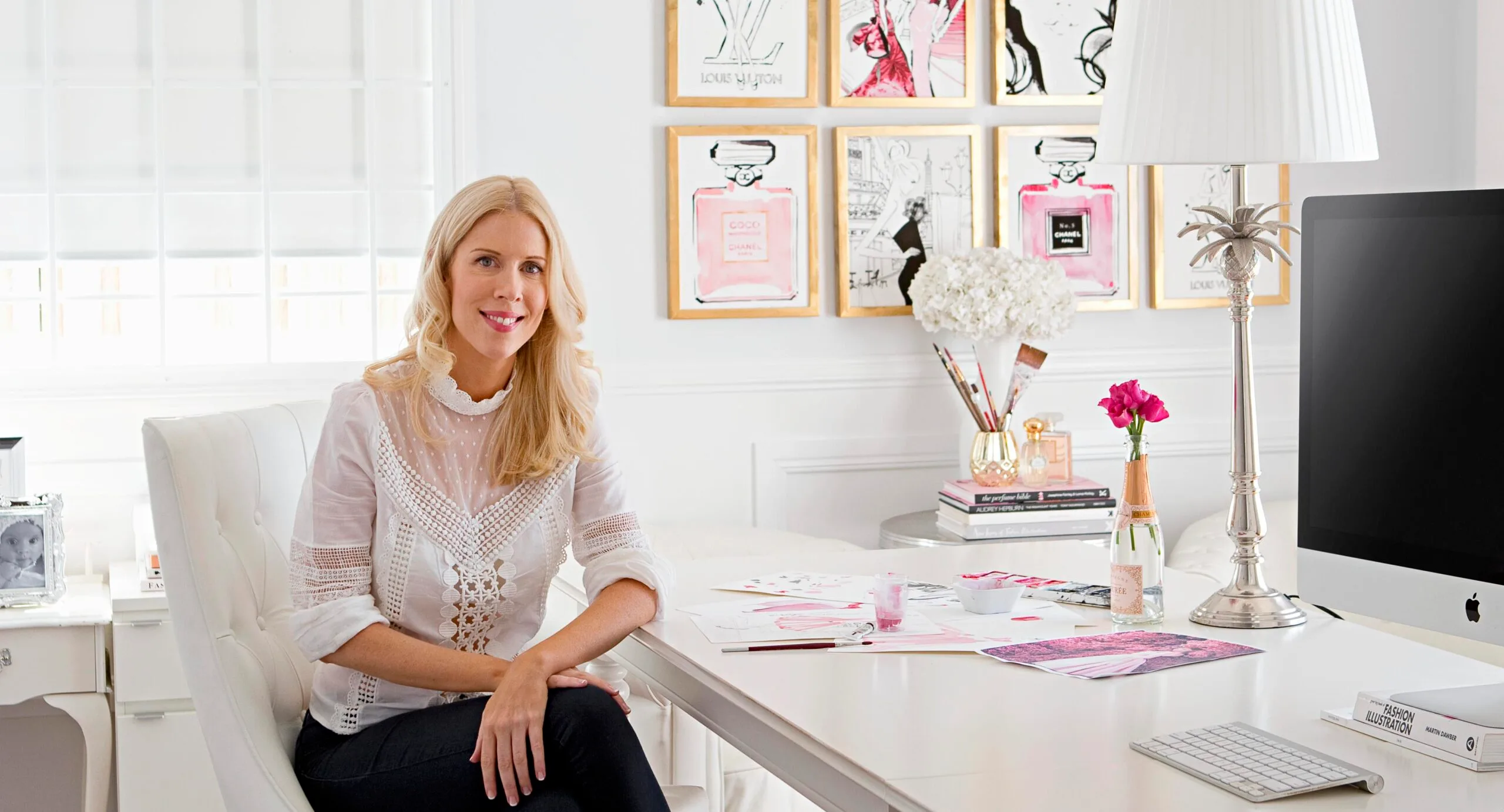 Blonde woman in a white blouse sits in a chic, white office with fashion artwork, flowers, and a desktop computer.
