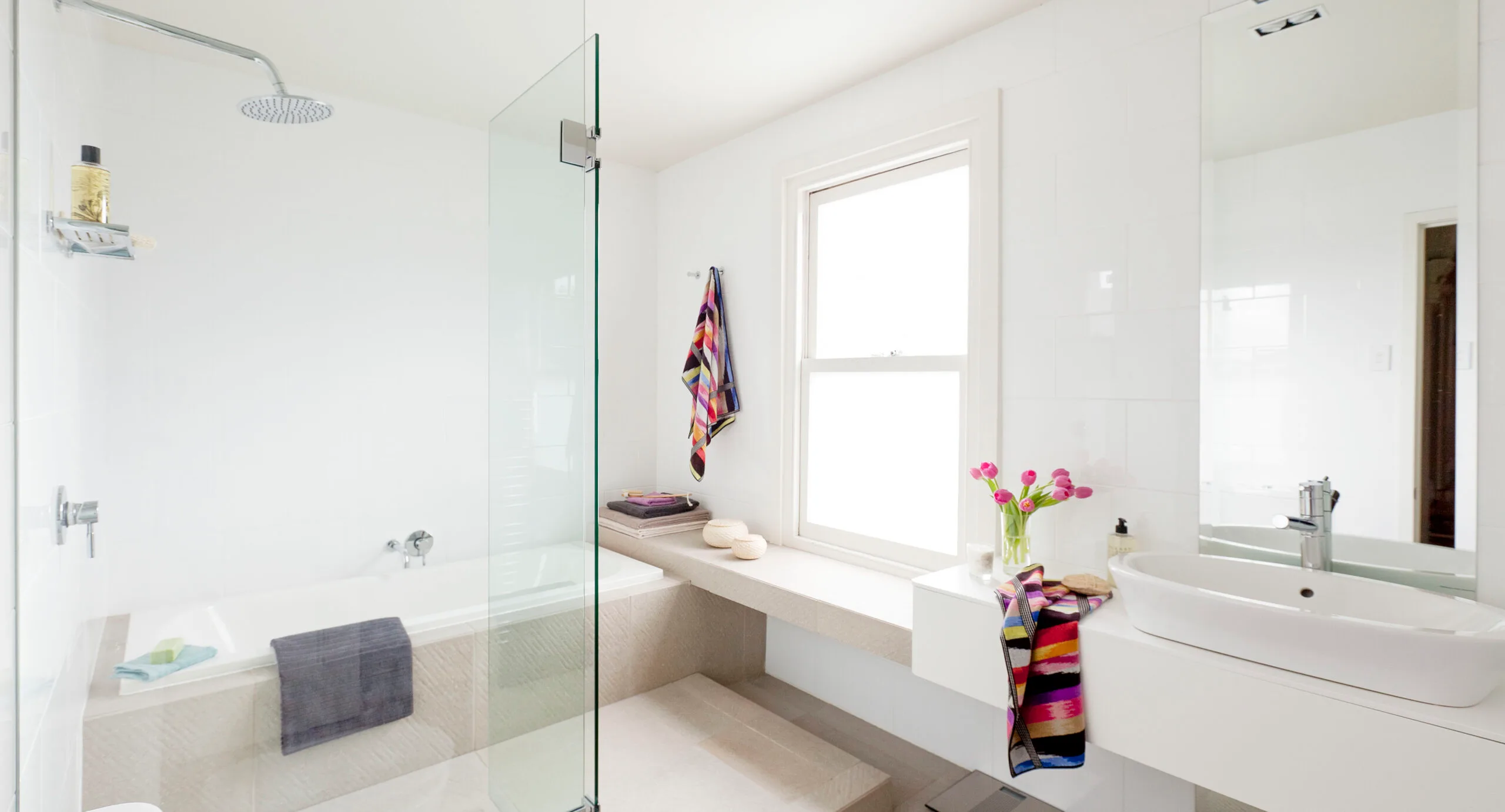 Bright modern bathroom with glass shower, bathtub, white walls, colorful towels, and a vase of pink flowers.