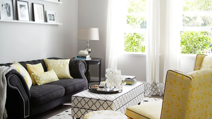 Modern living room with black sofa, yellow accents, patterned ottoman, and large window with sunlight streaming in.