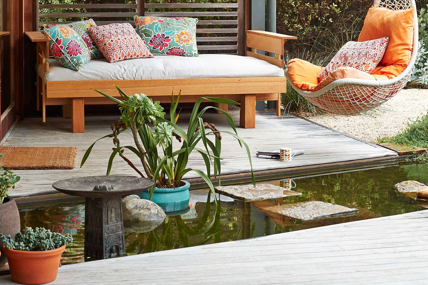 Cozy patio with colorful cushions on a bench and hanging chair, overlooking a small pond with stepping stones.