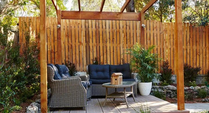 Outdoor patio with wicker furniture, wooden pergola, lush plants, and a birdhouse on the table.