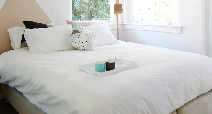 A neatly made bed with white bedding and decorative pillows, featuring a tray with two mugs on it by a window.
