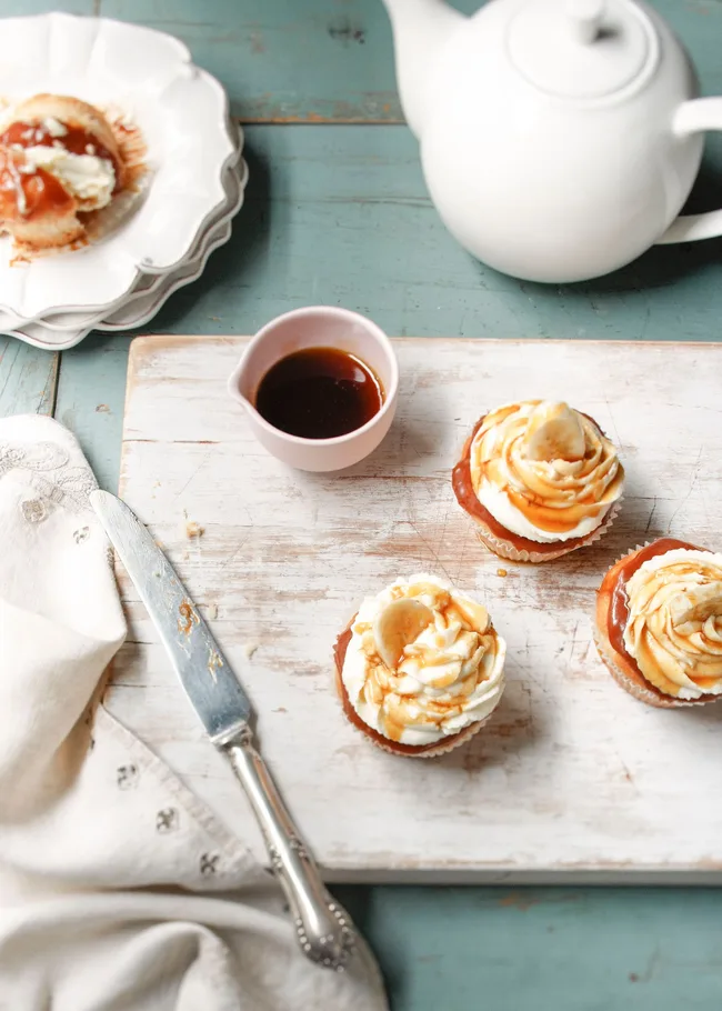 Cupcakes with cream and caramel drizzle on a wooden board, alongside a knife, white teapot, and small mug of sauce.