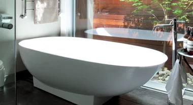 Freestanding white bathtub beside glass window with towel and toiletries nearby.