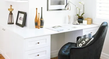 A modern white desk with a black chair, decor items, and a desk lamp in a bright room.