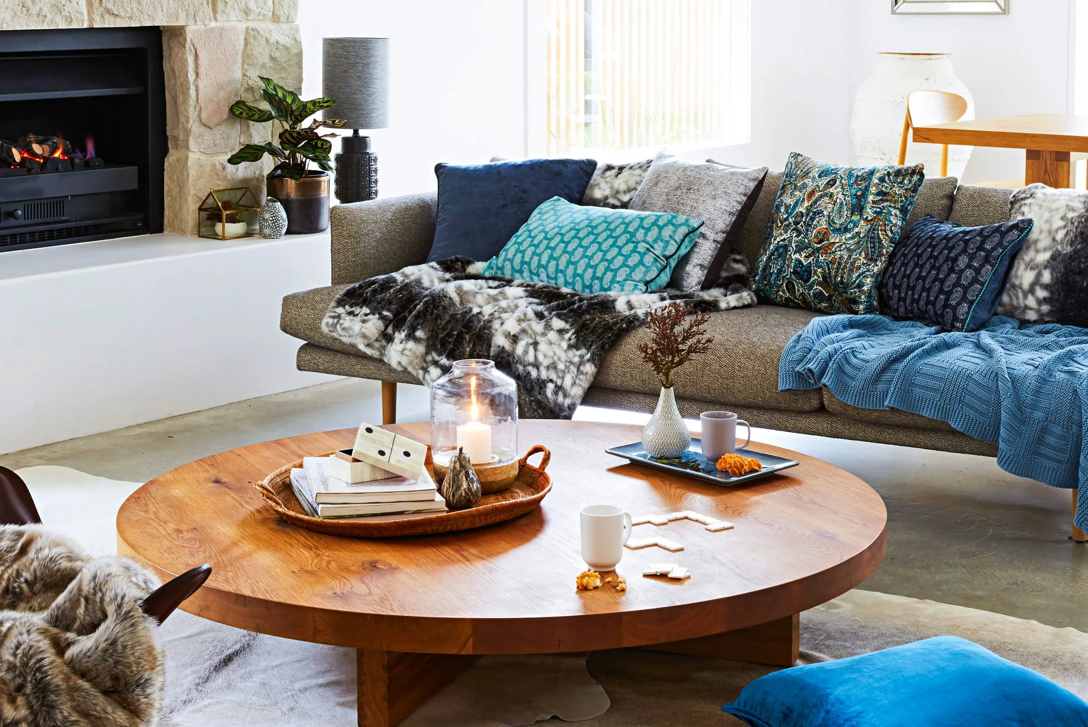 Cozy living room with gray sofa, colorful pillows, wooden coffee table, lit candle, and scattered magazines near a fireplace.