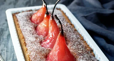 A rectangular tart with red poached pears standing upright, dusted with powdered sugar, on a dark textured background.