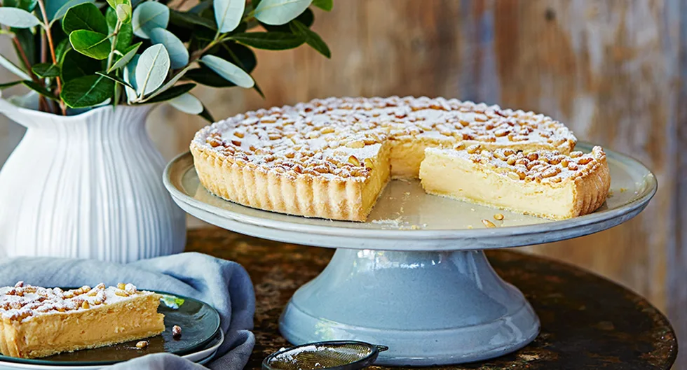 Custard tart with pine nuts dusted with powdered sugar on a cake stand, one slice cut and placed on a plate nearby.