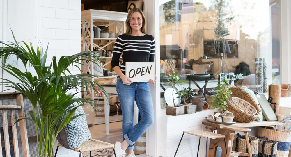 A woman stands smiling outside a store holding an "Open" sign, surrounded by home decor items.