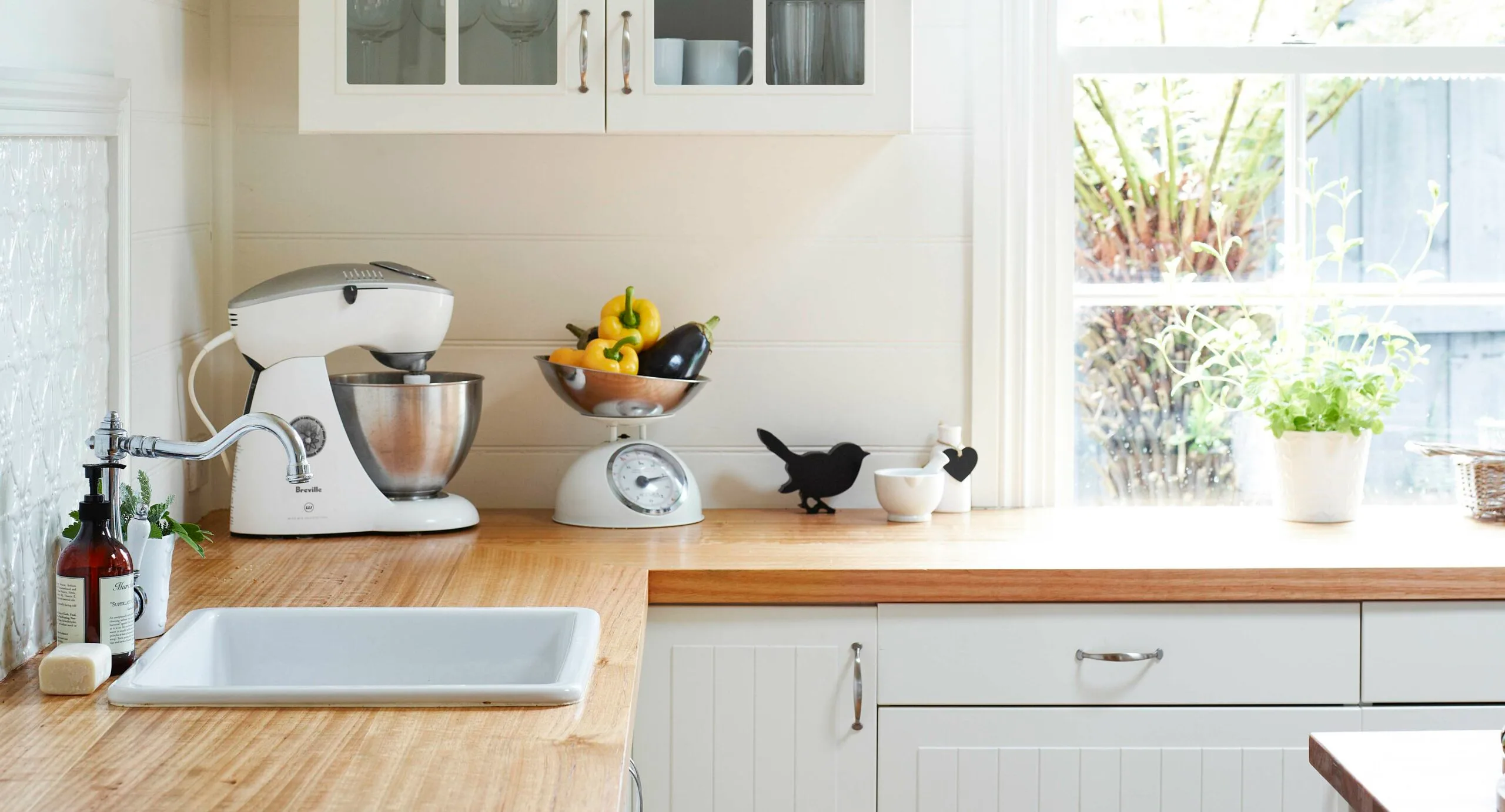 Bright kitchen countertop with mixer, scale holding vegetables, plant, and bird decor near a window.
