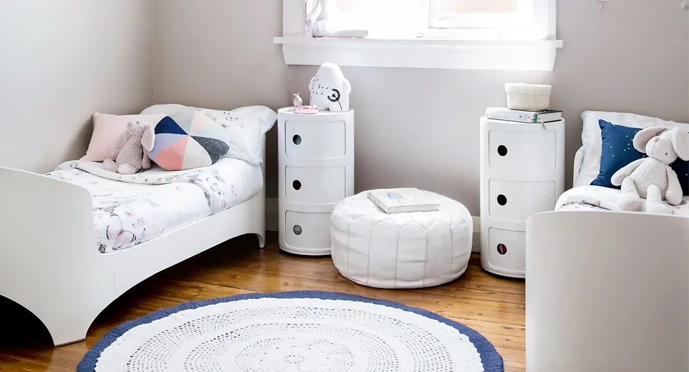 Cozy kids' room with two white beds, colorful pillows, two white dressers, a round ottoman, and a circular rug.