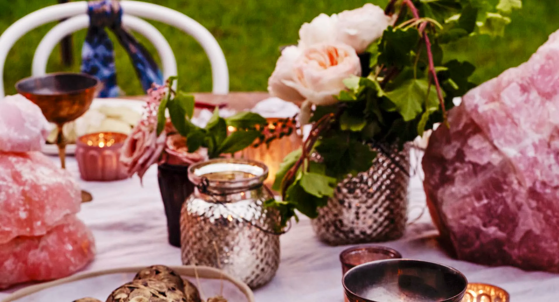 Outdoor table with pink crystals, flowers, metallic vases, and teacups set against a grassy background.