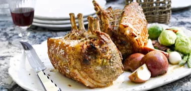 Roast crown of pork with herbs, served on a platter with red apples and Brussels sprouts.