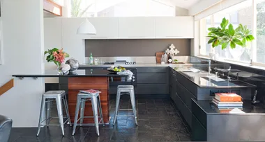 Modern kitchen with dark countertops, a small island with stools, decorative plants, and a large window view.
