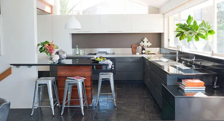 Modern kitchen with dark countertops, a small island with stools, decorative plants, and a large window view.