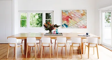 Modern dining room with a long wooden table, white chairs, colorful wall art, and a floral centerpiece.