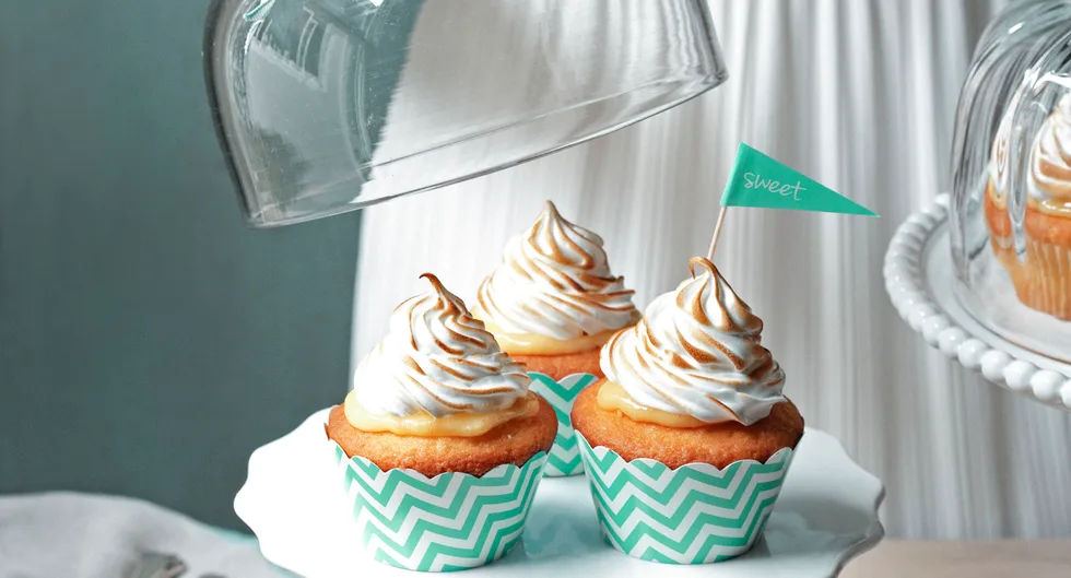Three cupcakes with toasted frosting on a white plate, one with a "sweet" flag, under a glass dome.