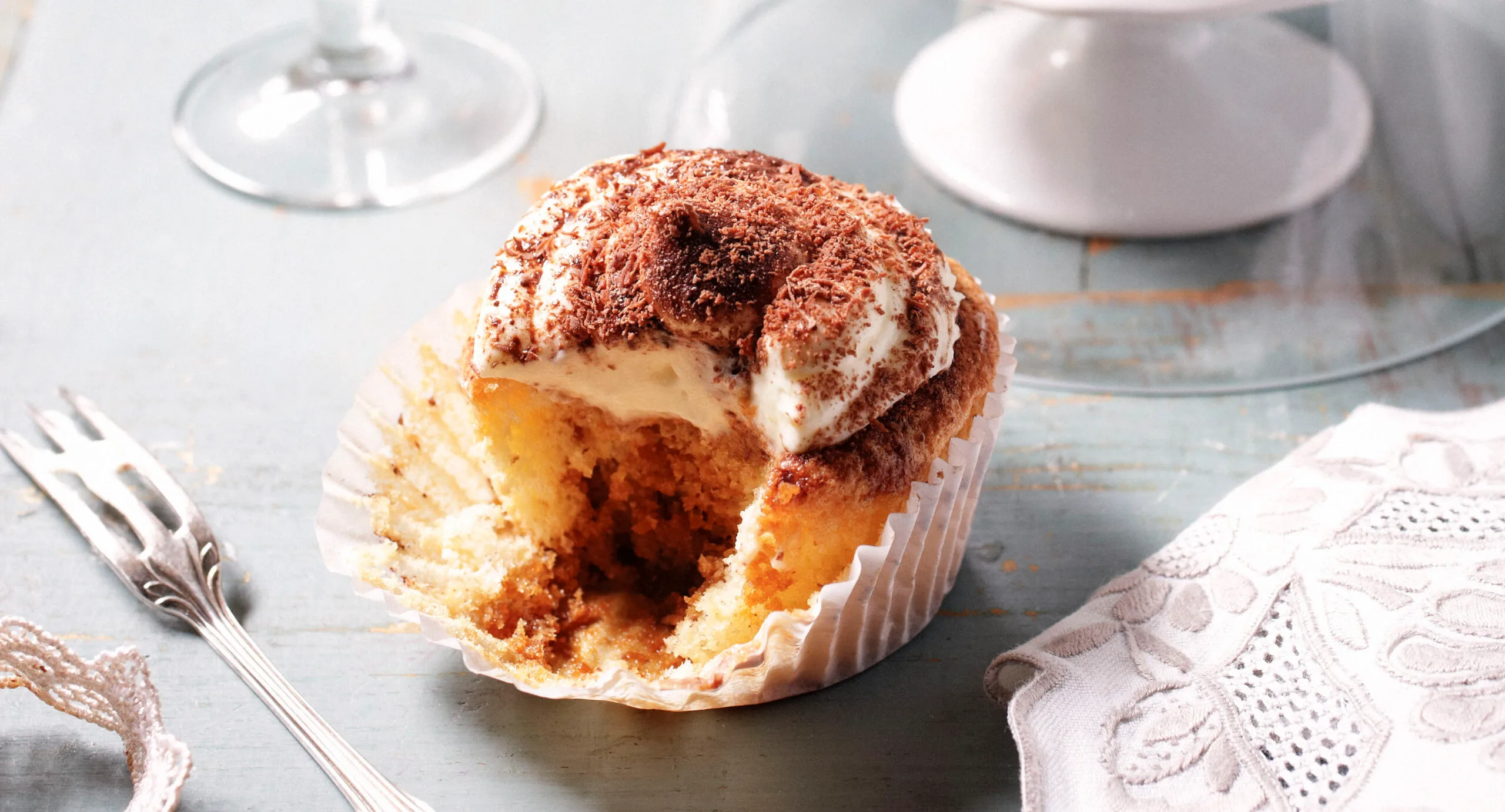 A tiramisu cupcake with a bite taken out, topped with cream and cocoa powder, on a light wooden surface.