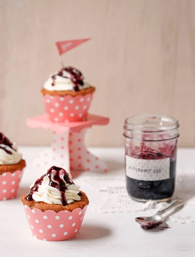 Cupcakes with white frosting and berry drizzle in pink polka dot wrappers, next to a jar of blueberry jam.