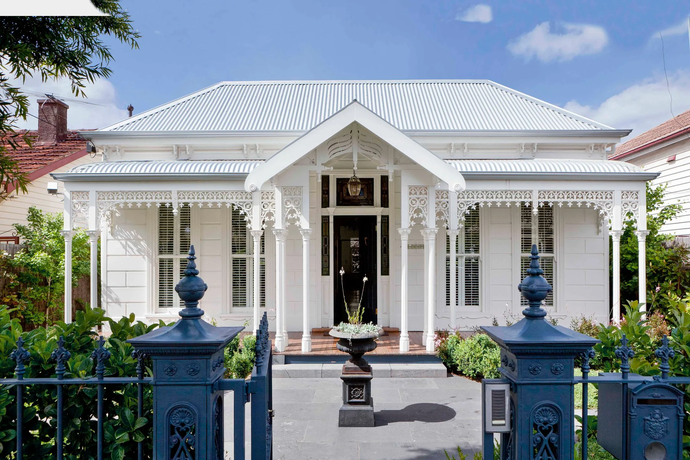 White Victorian-style house with ornate trim and blue fence, surrounded by greenery under a clear blue sky.