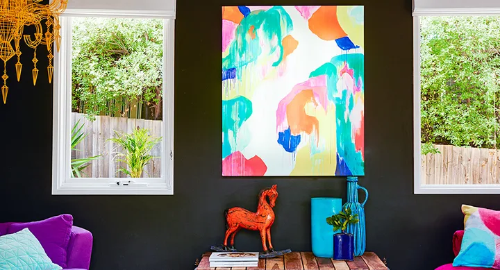 Bright living room with abstract colorful painting, purple sofa, and decorative items on a wooden table between two windows.