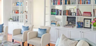 Living room with cream chairs, blue pillows, white shelves filled with books, decorative items, and pink flowers.