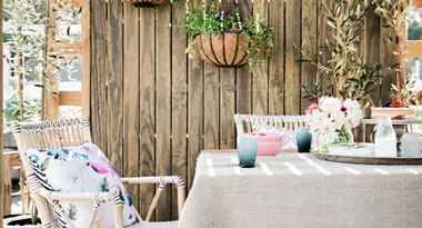 Outdoor patio scene with rattan chairs, floral cushion, table with flowers, cake, and milk, against a wooden fence backdrop.