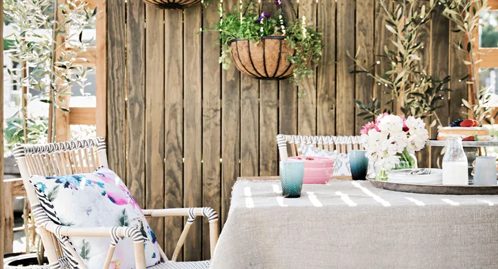 Outdoor patio scene with rattan chairs, floral cushion, table with flowers, cake, and milk, against a wooden fence backdrop.